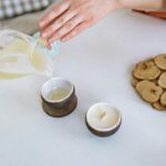 Close-up of hand pouring candle wax into rustic container, crafting essentials nearby.